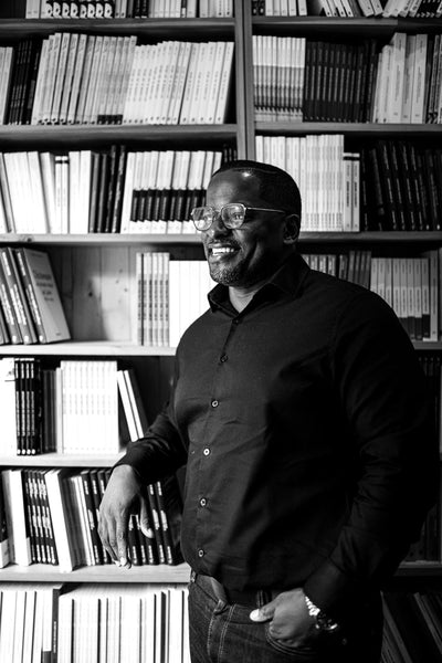 A man wearing glasses and a dark shirt stands smiling in front of shelves filled with neatly arranged books. The image is in black and white.