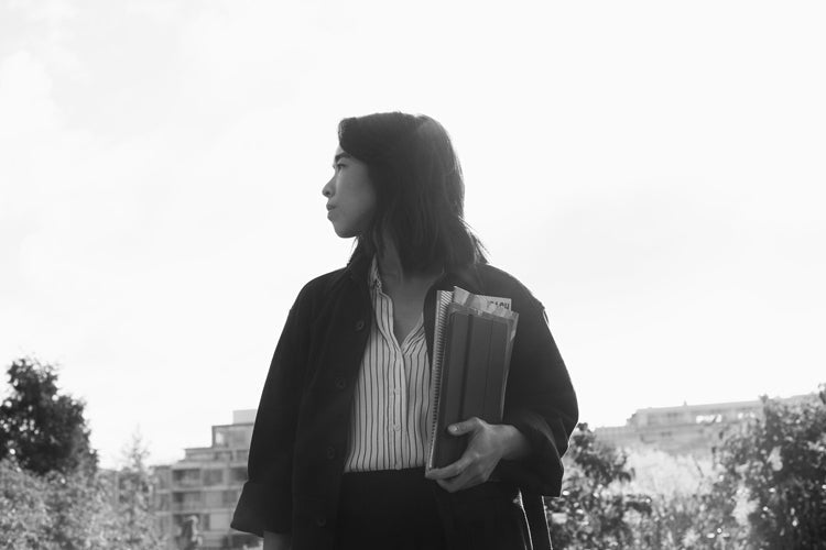 Black and white photo of a woman standing outdoors, holding folders and looking to her left. She is wearing a striped shirt and a dark jacket, with trees and blurred buildings in the background.