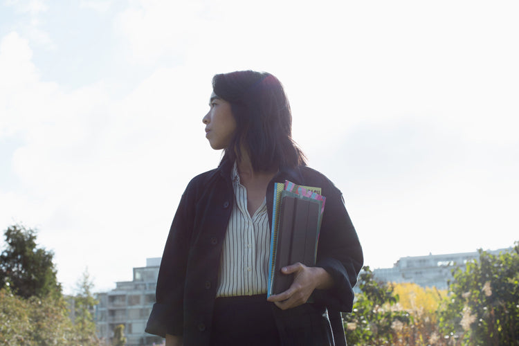 A woman stands outdoors holding folders and looking to the side. She wears a dark jacket over a striped shirt, with buildings and trees visible in the bright background.