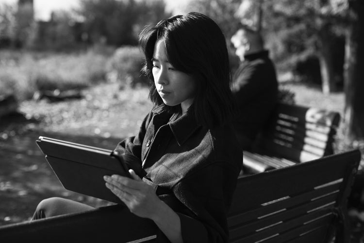 A woman sits on a park bench reading a tablet, with sunlight on her face. In the background, a man sits facing away on the same bench. The image is in black and white.