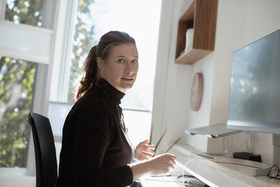 A woman with light brown hair in a ponytail sits at a desk in a bright, modern office, looking towards the camera. She wears a black turtleneck and is near a computer monitor with sunlight streaming in.