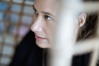 A close-up, slightly angled view of a woman‚Äôs face as she looks thoughtfully to the side. The image is soft-focused, with blurred foreground elements and a gentle, natural light.
