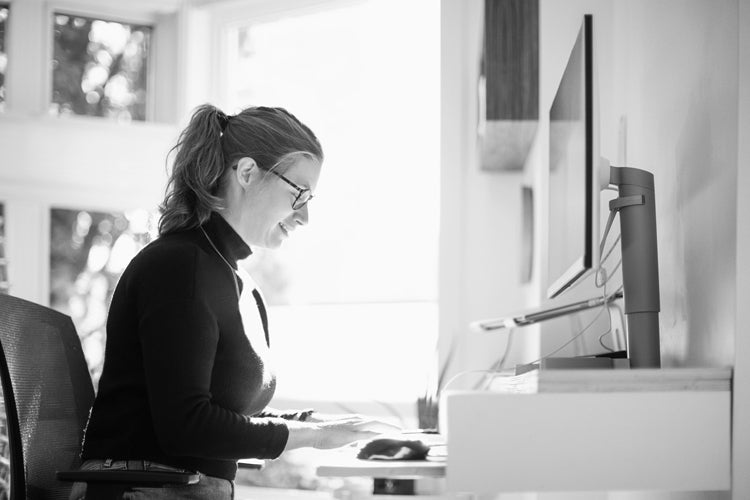 A woman wearing glasses and a turtleneck sweater sits at a desk, typing on a keyboard in front of a computer monitor, with sunlight streaming through large windows in the background.