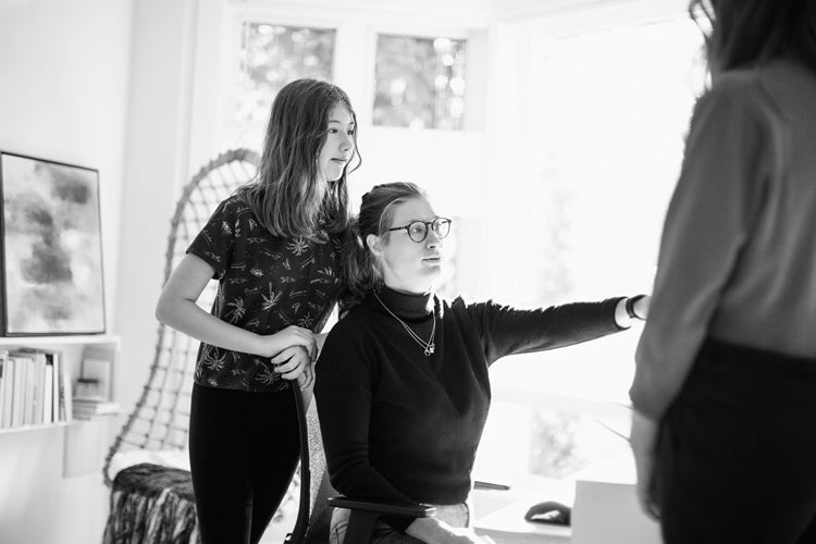 A woman sitting at a desk gestures toward another person while a girl stands next to her, all in a sunlit room with large windows and shelves in the background.