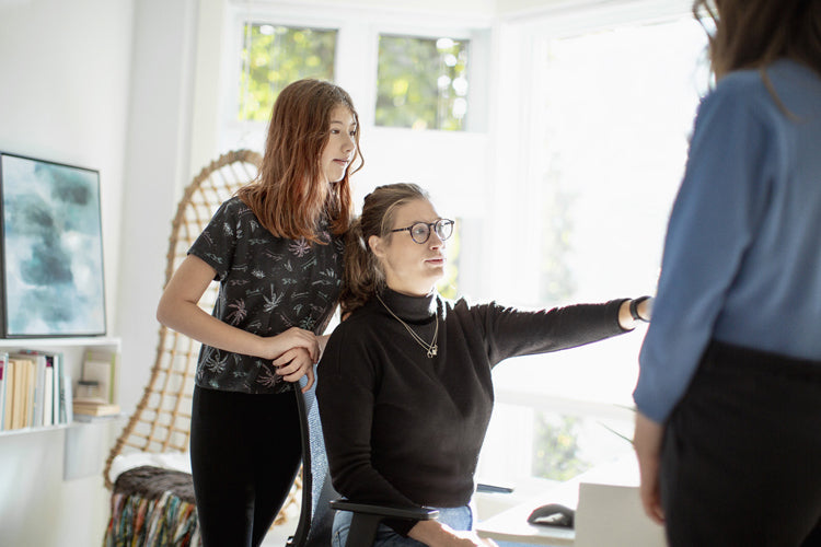 A woman seated at a desk points towards a standing person, while a young girl stands beside her. They are in a bright room with large windows and shelves.