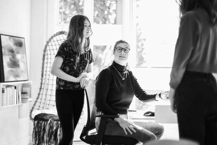 A woman sitting in an office chair talks to two standing people, one a teenage girl and the other an adult, in a bright, modern room with large windows and a bookshelf.