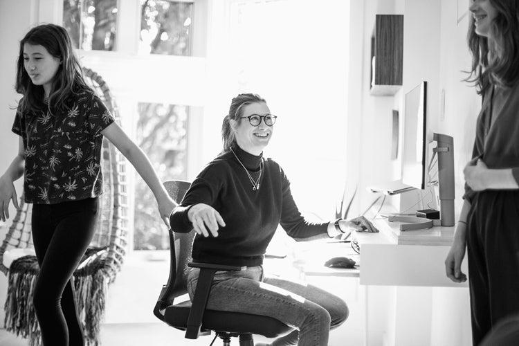 A woman wearing glasses sits and smiles at a desk with a computer, while two girls stand nearby. The scene is indoors, in a bright room with large windows.