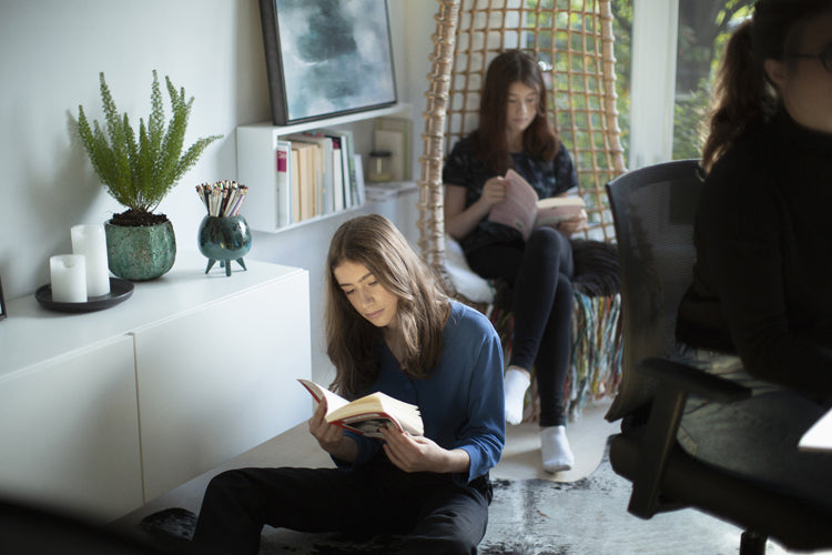 Three young people read books in a cozy, sunlit room. One sits on the floor, another in a hanging chair by the window, and the third at a desk. Shelves, plants, and candles decorate the modern, peaceful space.