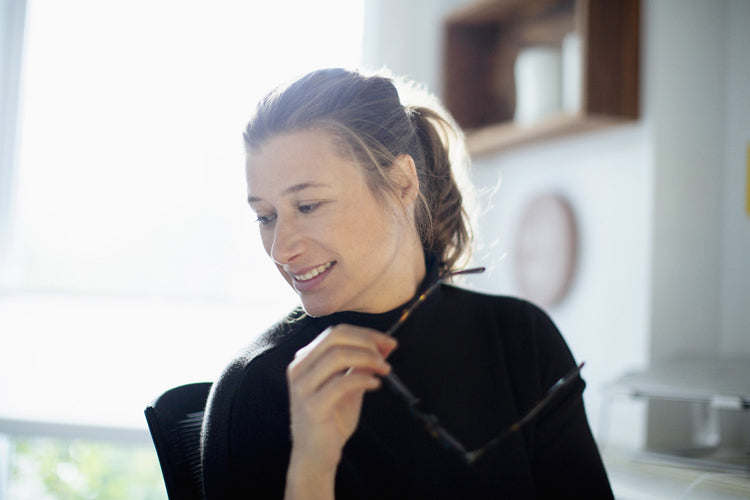 A woman with light brown hair in a ponytail sits indoors, holding a pair of glasses and smiling slightly while looking down. Sunlight streams through a window in the bright, modern room.