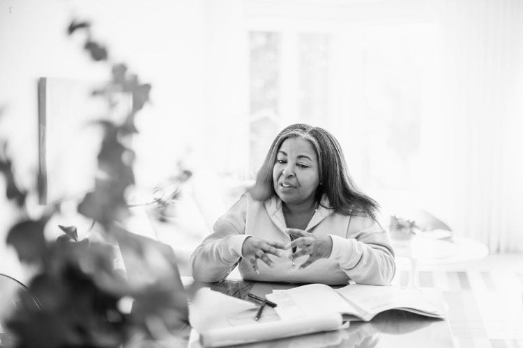 A woman sits at a table with open books and papers, gesturing with her hands as she talks, with natural light streaming in from a window behind her. The image is in black and white.