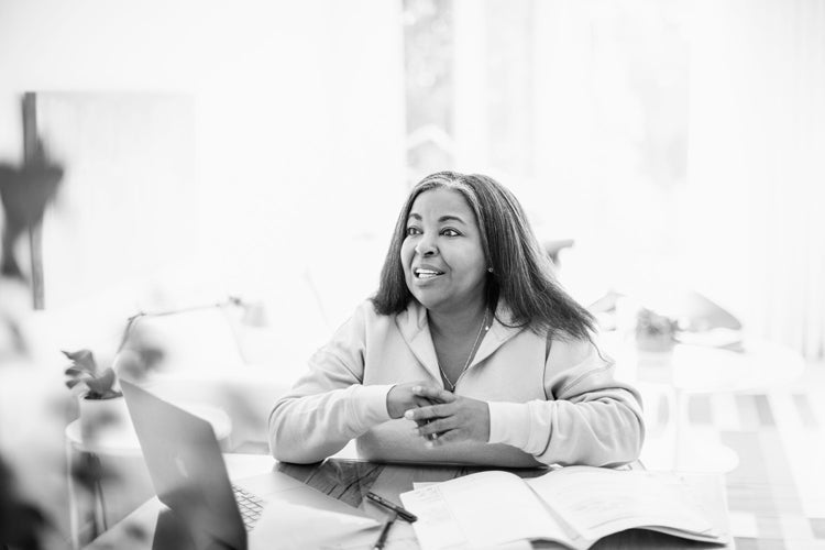 A woman sits at a table with a laptop and open books, smiling and looking up. The scene is bright and softly lit, creating a comfortable atmosphere for working or studying.