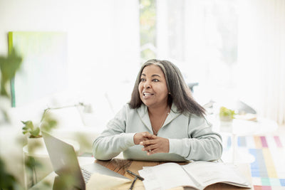 A woman sits at a table with a laptop, papers, and a pen, smiling and looking up. The background is bright and features a window and indoor plants.