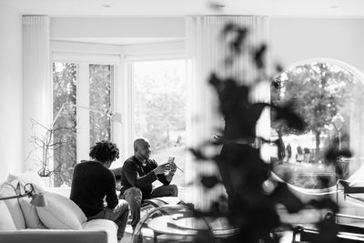 Two people sit and talk in a bright living room near large windows, with sunlight streaming in. The image is in black and white, and a leafy plant is out of focus in the foreground.