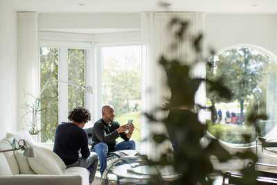 Two people sit and talk in a bright, modern living room with large windows, white furnishings, and green plants. Sunlight streams in, and trees are visible outside.