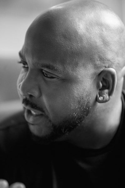 Black and white close-up photo of a man with a shaved head, a beard, and an earring, looking to the side with a serious expression.