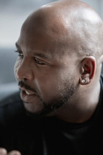 A close-up of a man with a shaved head and trimmed beard, wearing a stud earring and a dark shirt, looking to the side while speaking. The background is softly blurred.