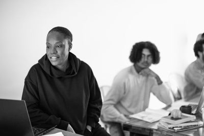 A person in a dark hoodie sits in front of a laptop, smiling, while two others sit at a table behind them with notebooks, one looking toward the camera. The scene is in black and white.