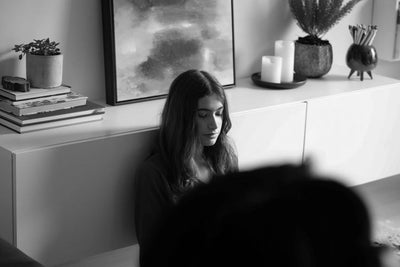 A woman with long hair sits on the floor against a cabinet, looking down thoughtfully. Behind her are candles, a potted plant, books, and a painting. The scene is in black and white.