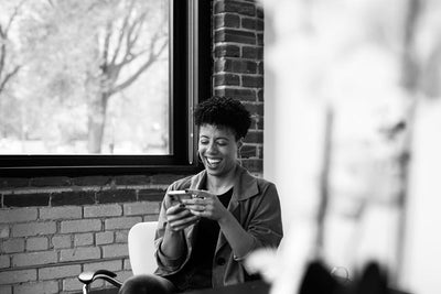 A person sitting in an office chair by a window, smiling and looking at their phone in a bright, modern workspace with exposed brick walls.