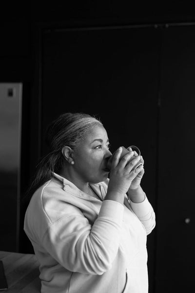 A woman in a light-colored sweatshirt sips from a mug, looking thoughtfully to the side. The background is dark, creating a contrast with her clothing. The image is in black and white.