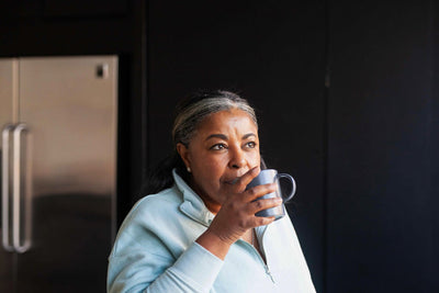 A woman with gray-streaked hair, wearing a light blue sweater, stands indoors near a refrigerator and drinks from a gray mug, looking thoughtfully into the distance.