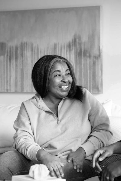 A woman with long hair smiles warmly while sitting on a couch, wearing a light-colored hoodie. One hand rests on her knee, and she‚Äôs engaged in conversation. The background features a large, abstract painting.