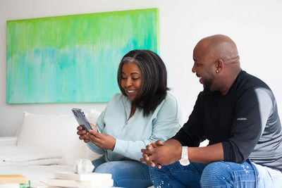 A woman smiling while looking at her phone, sitting on a couch next to a man who is looking at her, both appearing happy and relaxed in a bright room with a green abstract painting on the wall.