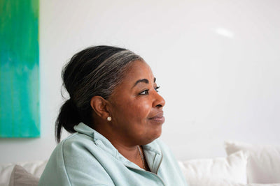 A woman with gray-streaked hair tied back is sitting indoors, wearing a light blue top and small earrings, looking contentedly to the side. A white wall and a green painting are in the background.