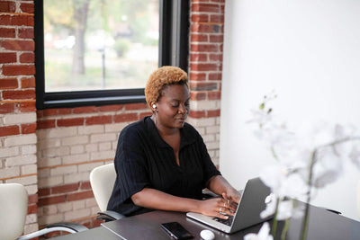 A woman with short curly hair sits at a table using a laptop in a bright, modern office with brick walls and large windows. A smartphone and white flowers are on the table.