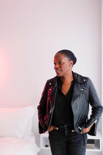 A woman wearing a black leather jacket and black pants stands with her hands in her pockets in a minimal, white room next to a bed with white sheets. She looks to the side and appears relaxed.