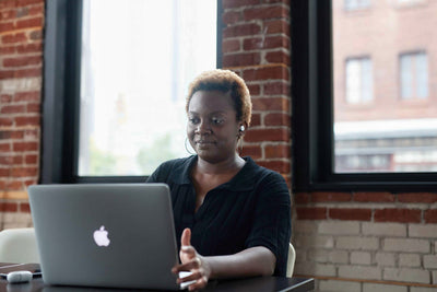 Woman sits at a desk working on a laptop in a modern office with exposed brick walls and large windows, looking at the screen with a focused expression.