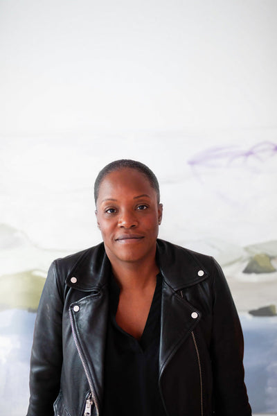 A person with short hair wearing a black leather jacket and black shirt stands in front of a light-colored, abstract background, looking directly at the camera with a neutral expression.