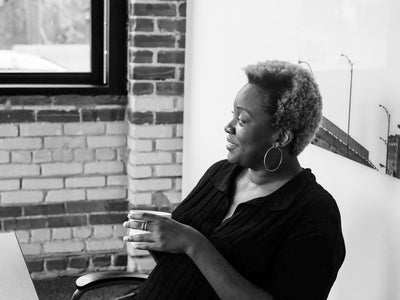 Black-and-white photo of a woman with short curly hair, wearing hoop earrings and a dark top, smiling while holding a cup. She is seated indoors near a window and exposed brick wall.