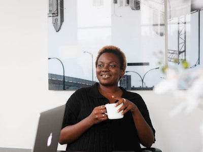 A person with short hair is smiling while holding a white mug, seated at a desk with a laptop. A modern cityscape photo hangs on the wall in the background.