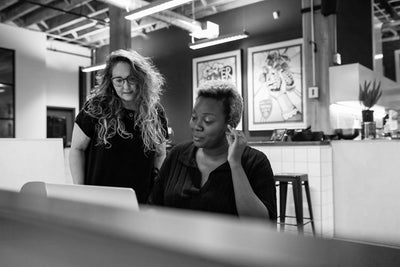 Two women in an office setting look at a laptop together. One woman is seated, touching her ear, while the other stands beside her, both appearing focused. The workspace is modern with artwork on the wall behind them.