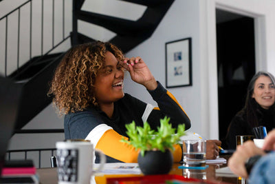 A woman with curly hair laughs while sitting at a table with papers, a coffee mug, a plant, and a glass of water. Another woman is smiling in the background near a staircase.