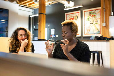 Two women sit at a desk in a modern office, engaged in conversation. One woman gestures with her hands while the other, smiling, watches. Colorful artwork is on the wall behind them.