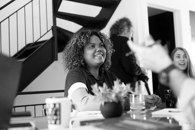 A woman with curly hair sits at a table, smiling during a meeting. Another woman and a blurred person are in the background near a staircase. Papers and a potted plant are on the table. The scene is in black and white.