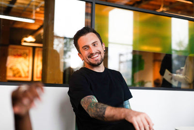 A man with tattoos on his arms, wearing a black t-shirt, sits and smiles in a modern office with glass walls and colorful decor.