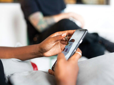 Close-up of a person holding a smartphone and using apps, with another person sitting in the blurred background. The focus is on the hands and phone.