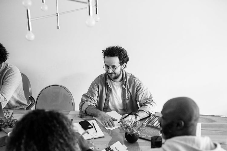 Black and white photo of a man with glasses sitting at a table with papers, surrounded by three other people, having a meeting or discussion in a modern office setting.