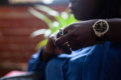 A person wearing a blue shirt, a large ring, a gold ring, and a wristwatch is adjusting their fingers. The background is blurred with hints of a plant and a brick wall.