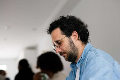 A man with curly dark hair, glasses, and a beard, wearing a light denim shirt, sits indoors looking down thoughtfully. The background is softly blurred with people and neutral colors.