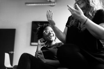 Two women sit indoors, laughing and talking. One woman with curly hair and glasses smiles brightly, while the other gestures animatedly with her hands. The scene is casual and joyful, captured in black and white.