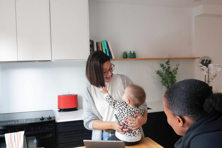 A woman holding a baby smiles while interacting with another person in a modern kitchen. The baby reaches out toward the woman, and kitchen shelves and appliances are visible in the background.