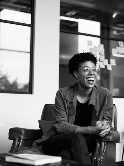 A person with short curly hair and glasses sits in an armchair, smiling and laughing. They wear a jacket over a t-shirt. Behind them are windows and a wall with sticky notes. The image is black and white.