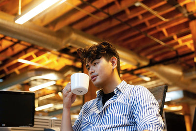 A person with wavy hair, wearing a striped shirt, sits in an office holding a white mug. The background shows exposed wooden beams and ductwork with warm overhead lighting.