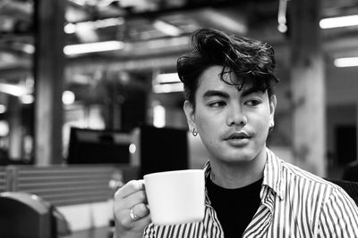 A person with short, wavy hair holds a mug and looks to the side, sitting in an office setting. The image is black and white, and the individual wears a striped shirt and earrings.
