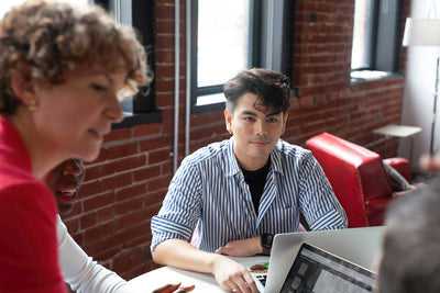 A group of people sit around a table in a modern office with brick walls, focusing on a discussion. One person in a striped shirt looks attentive, with laptops and papers on the table.