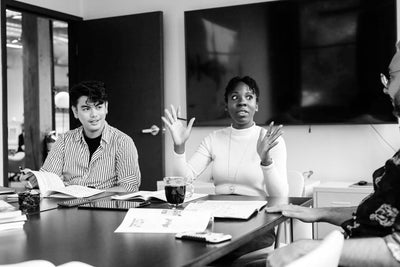 Three people sit around a conference table with open books and notebooks. A woman in a white top gestures while speaking, as the others listen. The setting appears to be a collaborative office meeting.